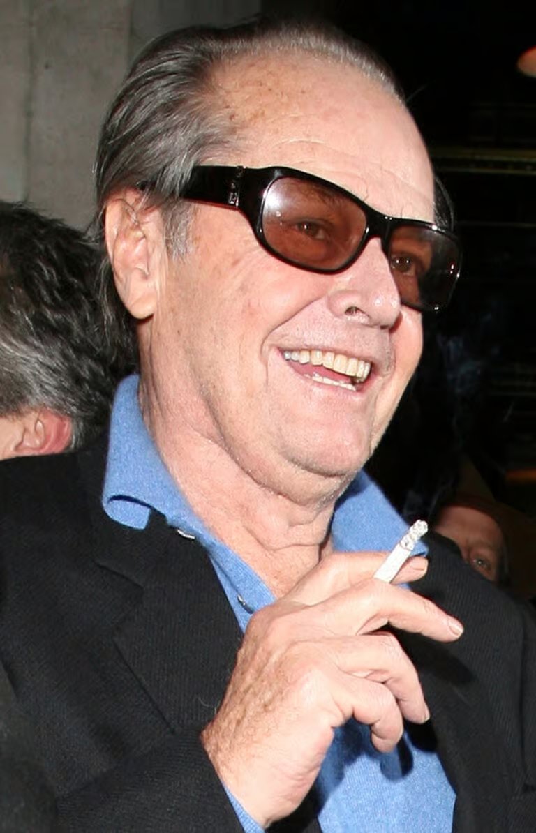 Jack Nicholson, con gafas de sol y camisa azul, sonríe mientras sostiene un cigarrillo